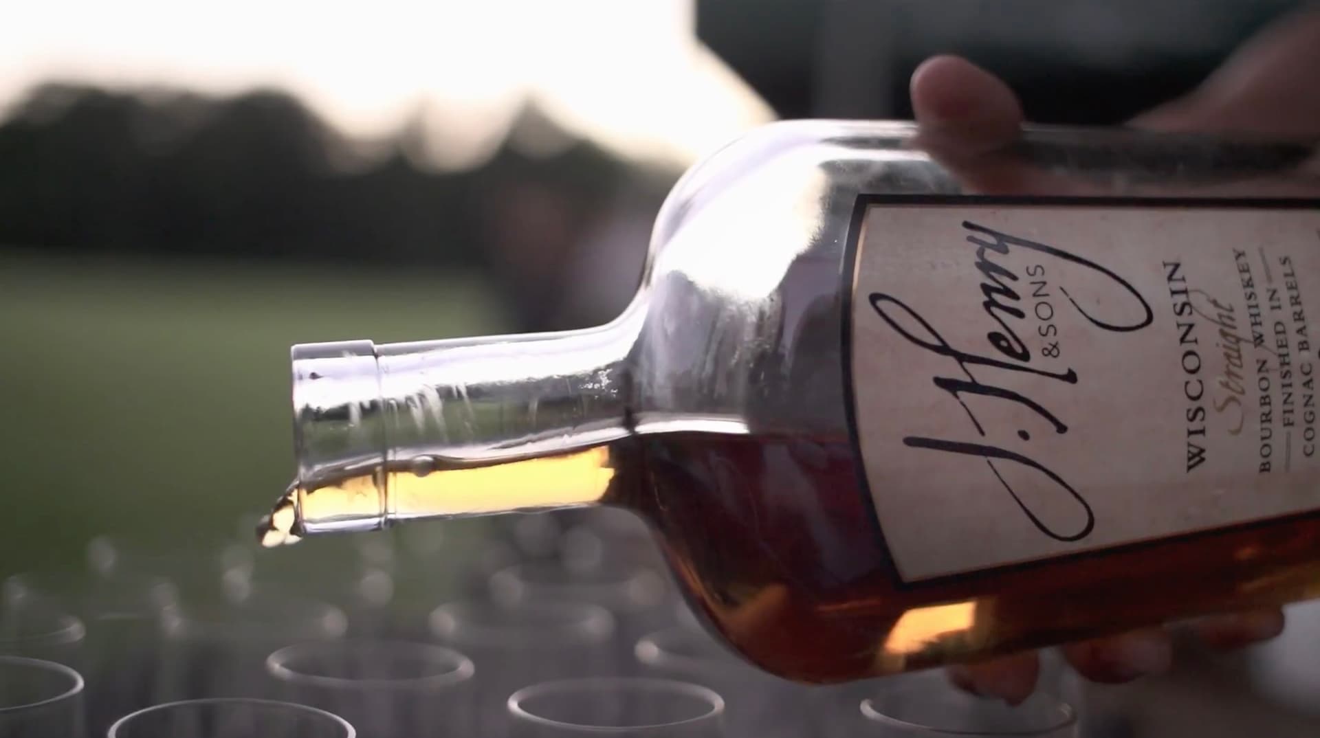 A hand pouring J. Henry & Sons Wisconsin Straight Bourbon, finished in cognac barrels, into a row of glasses
